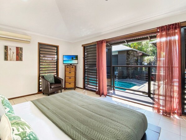 Un dormitorio con cama, una silla, televisor y puerta corrediza de vidrio que da a una piscina.