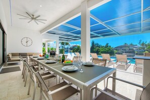 A spacious outdoor dining area with a long table, chairs, and a view of a pool and tropical landscape.