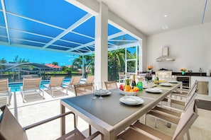 Une cuisine moderne dotée d’un grand îlot central, d’un coin repas offrant une vue sur la piscine et d’une terrasse avec des fauteuils de jardin.