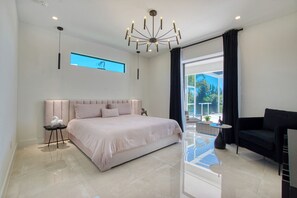A modern bedroom with a large bed, a black armchair, a small table, and a view of the outdoors.