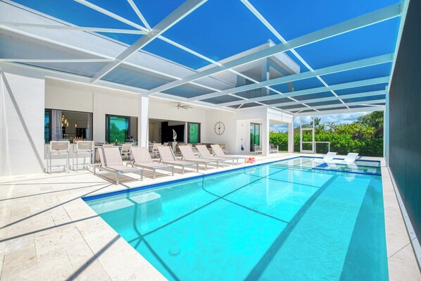 A modern indoor pool area with clear blue water, white lounge chairs, and a glass ceiling structure.