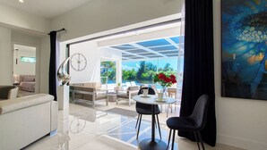 A modern living room with a sofa, a coffee table, and a dining area with chairs. There is a large window with a view of greenery and a pool. - VRCC Villa Sogno - modern-day getaway (Cape Coral)