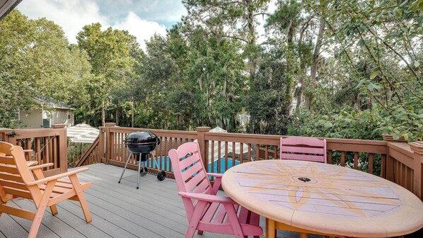 A wooden deck with a round table and chairs, a grill, and a view of a swimming pool and trees.