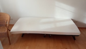 Ein Einzelbett mit weißer Matratze in einem Zimmer mit Holzboden und einem Korbstuhl.