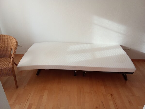 A single bed with a white mattress in a room with wooden flooring and a wicker chair.