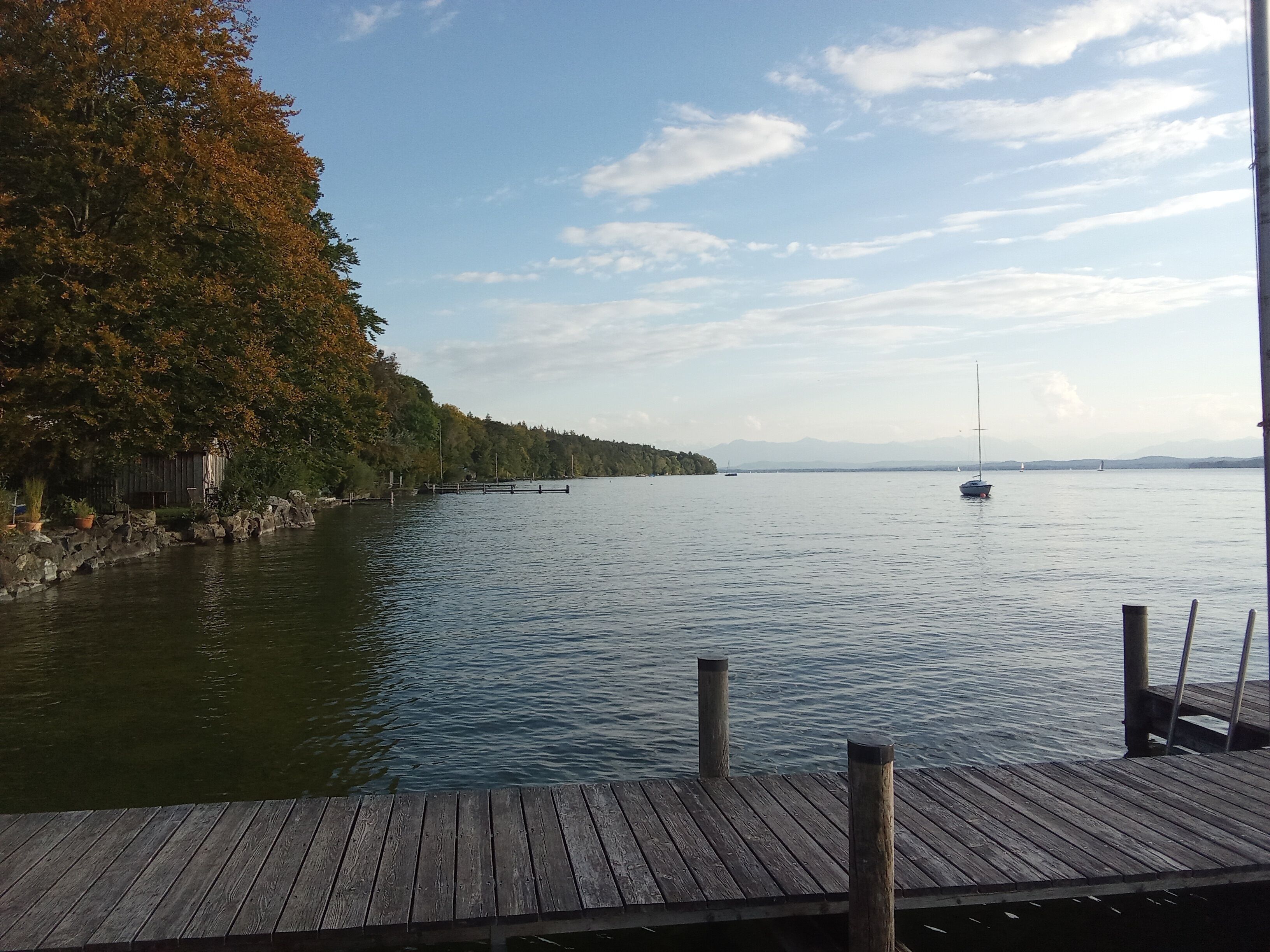 Eine Holzsteg, der sich in einen ruhigen See erstreckt, mit einem Segelboot in der Ferne.