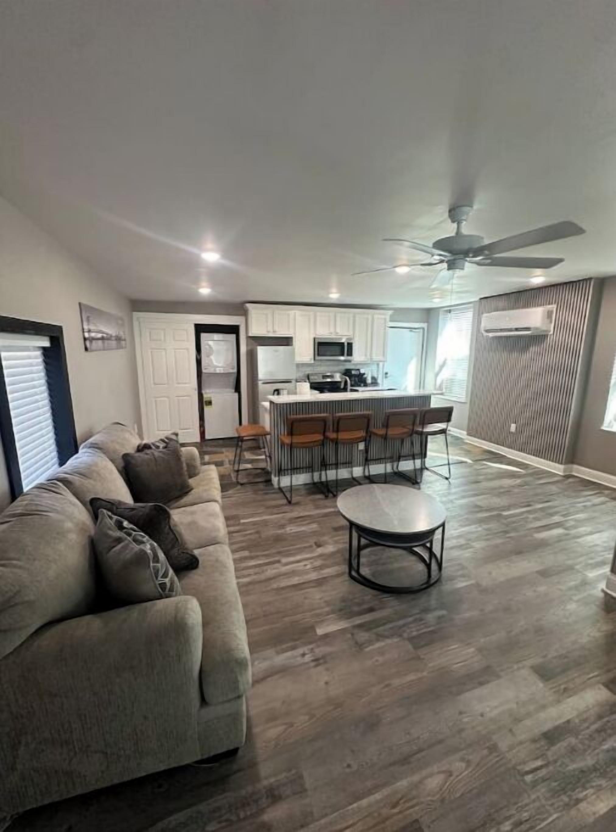 A spacious living room with a large gray sofa, a round coffee table, and a kitchen area with white cabinets and a microwave.