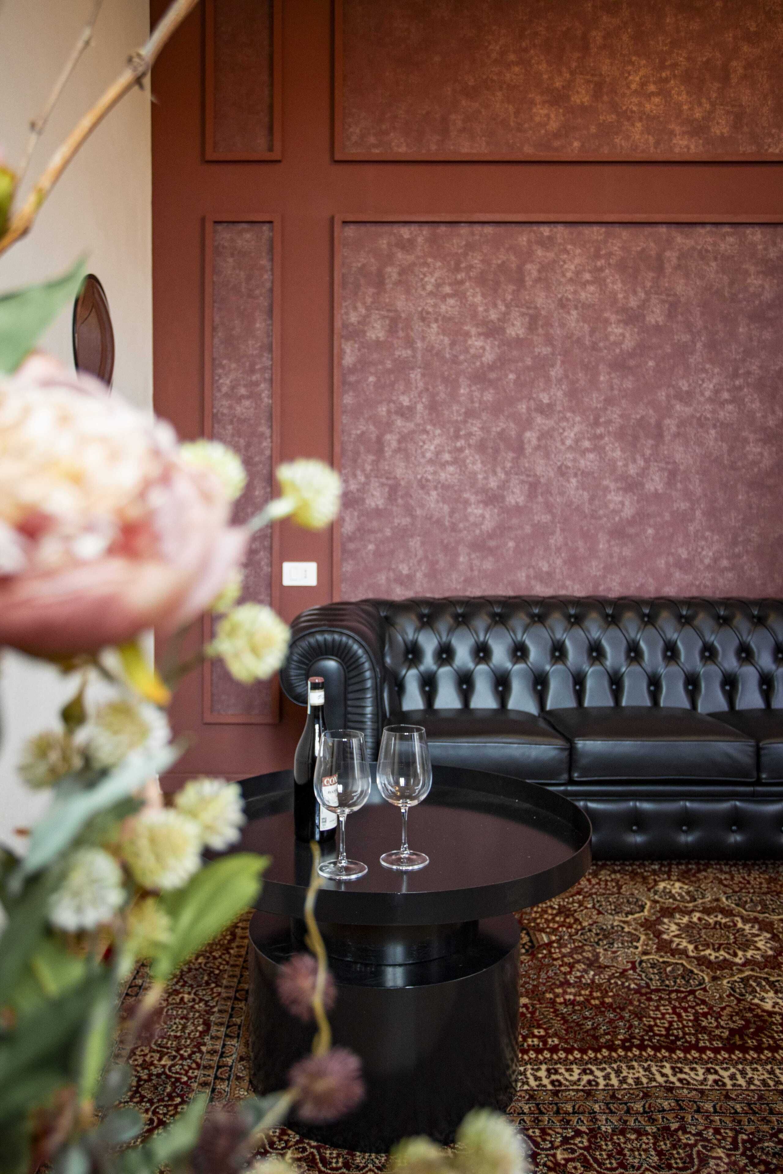 A room with a black leather sofa, a round black table with two wine glasses, and a floral arrangement.