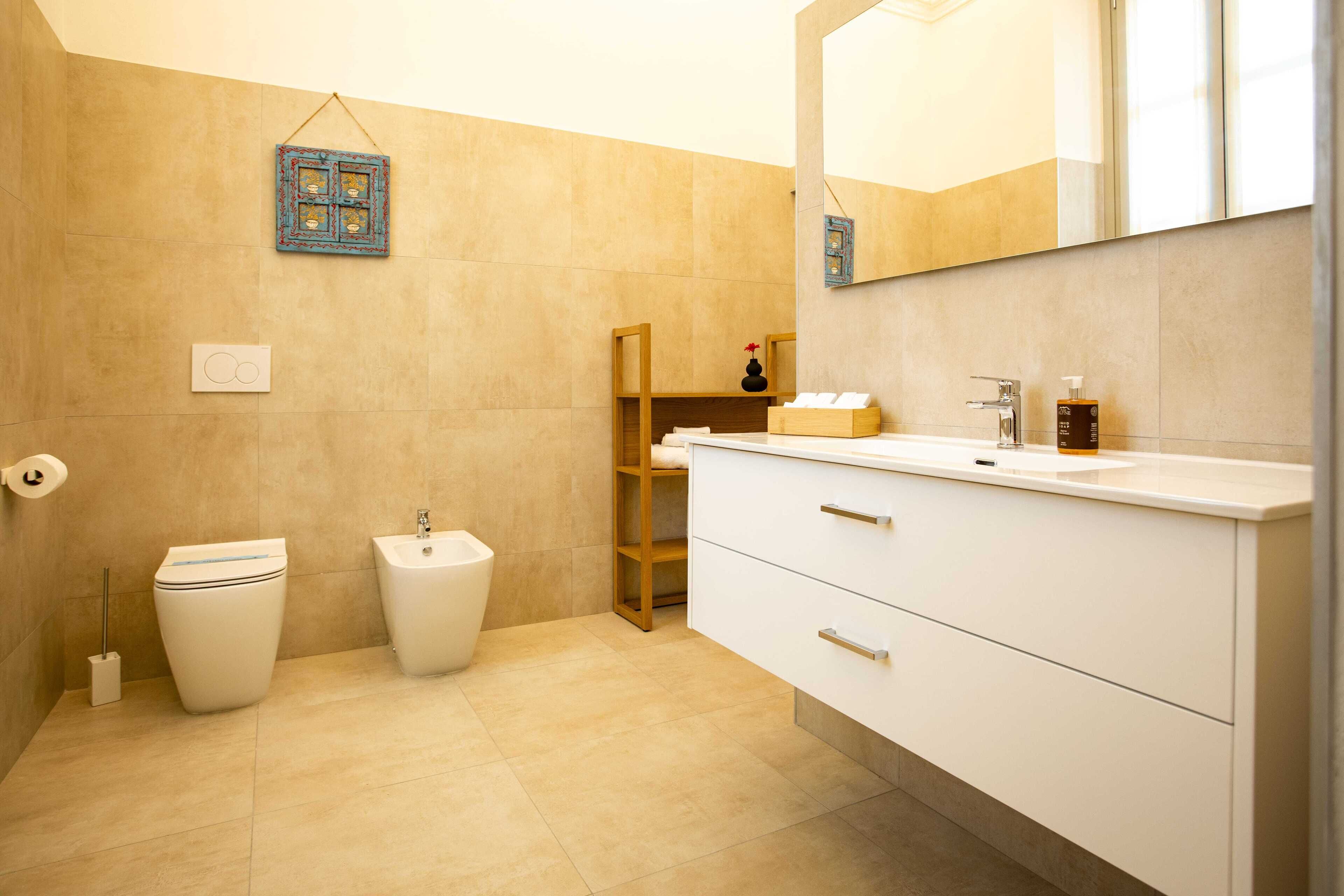 A modern bathroom with a toilet, bidet, vanity with a mirror, and a shelf with toiletries.