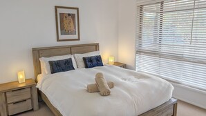 A neatly made bed with a rolled towel, two bedside lamps, and a framed artwork above the headboard. - Milo's @ the Bay by Experience Jervis Bay (Vincentia)