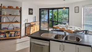 A modern kitchen with a large window, a dining area, and a bookshelf filled with books. - Milo's @ the Bay by Experience Jervis Bay (Vincentia)