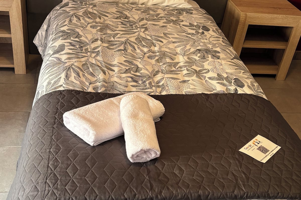A neatly made bed with a floral-patterned duvet, a quilted brown bedspread, and two rolled towels. There are wooden nightstands on either side of the bed, and a small table with a card on it.