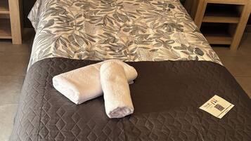 A neatly made bed with a floral-patterned duvet, a quilted brown bedspread, and two rolled towels. There are wooden nightstands on either side of the bed, and a small table with a card on it.