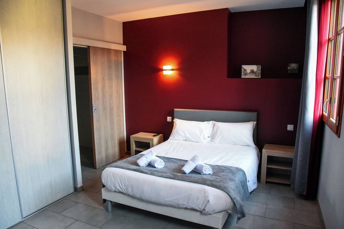 A neatly made bed with white linens and two rolled towels, a bedside table, a wall-mounted light, and a small picture frame on the wall.