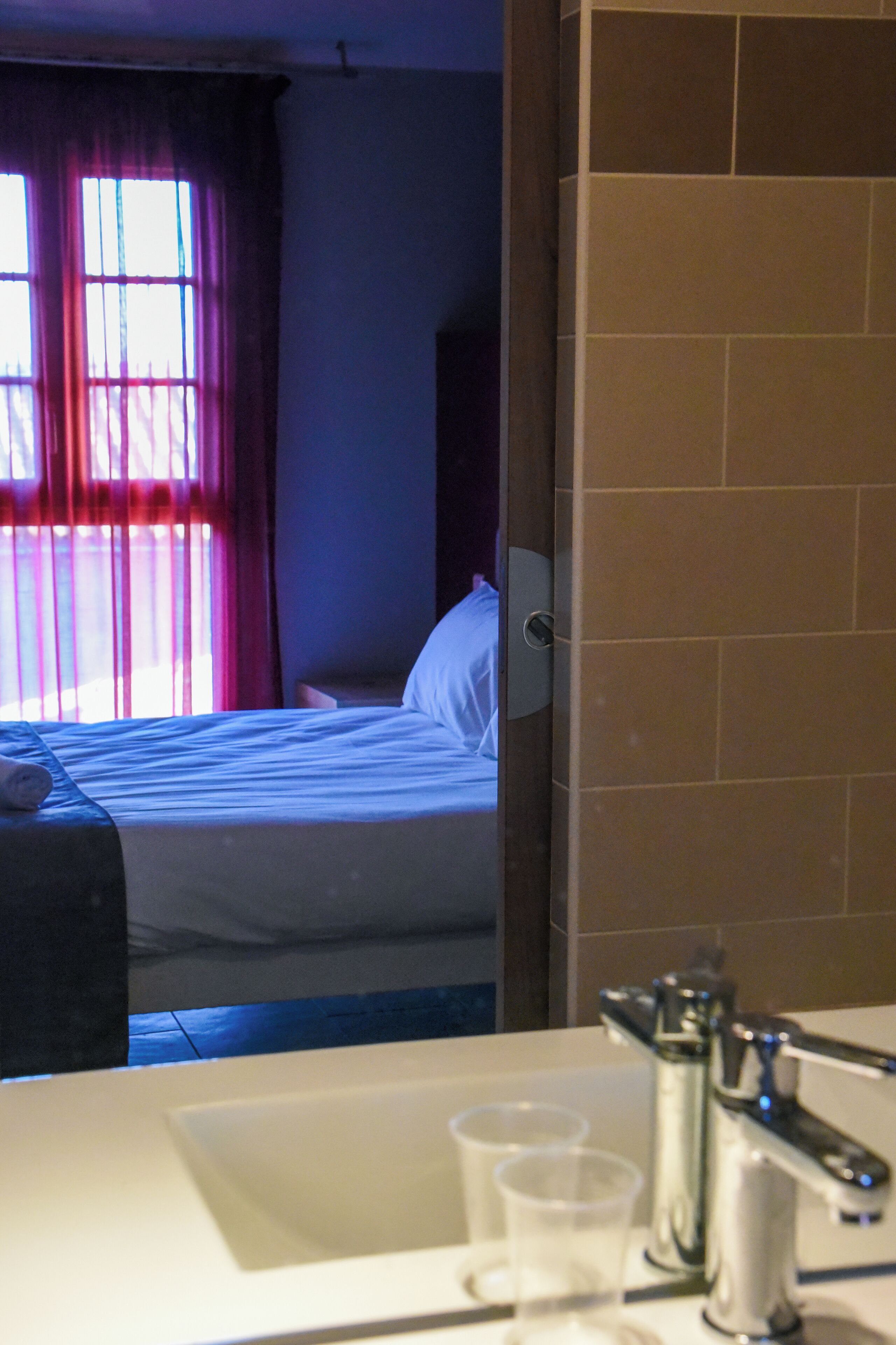 Ein Hotelzimmer mit einem Bett, einem Fenster mit roten Vorhängen sowie einem Badezimmer mit Waschbecken und zwei Gläsern.