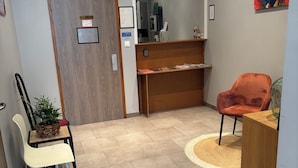 Un hall d’hôtel comprenant un comptoir d’accueil, un espace d’attente avec une chaise et une petite table, une plante en pot et un tableau accroché au mur.