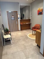 Zona de recepción de un hotel con un escritorio de madera, mostrador de recepción, área de espera con una silla, una planta en maceta y una cámara de seguridad instalada en la pared.