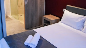 Une chambre d’hôtel comprenant un lit, une table de chevet, une lampe fixée au mur et une porte coulissante donnant accès à une cabine de douche.