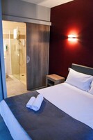 Habitación de hotel con una cama, una mesita de noche, una lámpara de pared y una puerta corredera que da a la zona de la ducha.
