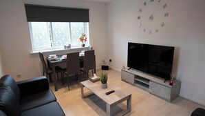 A modern living room with a sofa, a coffee table, a dining area with chairs and a table, a TV, and a clock on the wall. - Beckton Docklands - 1 Bedroom - Sleeps 4 - Wifi (London)