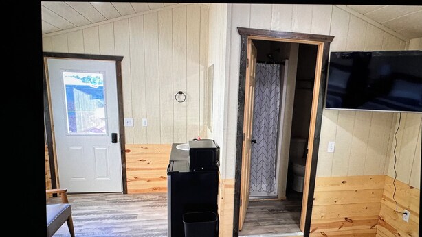 A room with wooden walls, a door with a window, a sink, a trash bin, and a television mounted on the wall. - O’Neill Ventures’s Queen Cabin with WIFI (Martin)
