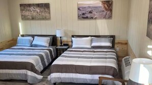 A room with two beds, a small table, and a chair. There are two wall-mounted lamps and a heater. - O’Neill Ventures’s Queen Cabin with WIFI (Martin)