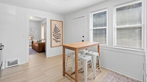 A modern living room with a wooden table and white bar stools, a large window with blinds, and a door leading to another room. - Comfortable 2- Bedroom House In Cleveland 1330 (Cleveland)