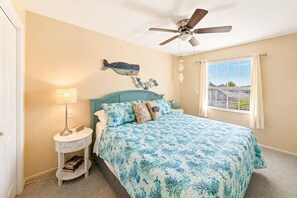 A bedroom with a bed, a nightstand, a lamp, a ceiling fan, a window with curtains, and a wall decoration. - Melbourne Beach Oceanfront Apartment Unit #2 - Ocean Views (Melbourne Beach)