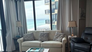 Un soggiorno con un divano color beige, una poltrona in pelle nera, un tavolino in legno e una ventola a soffitto. Grandi finestre offrono vista sulla spiaggia e su un edificio con balconi.