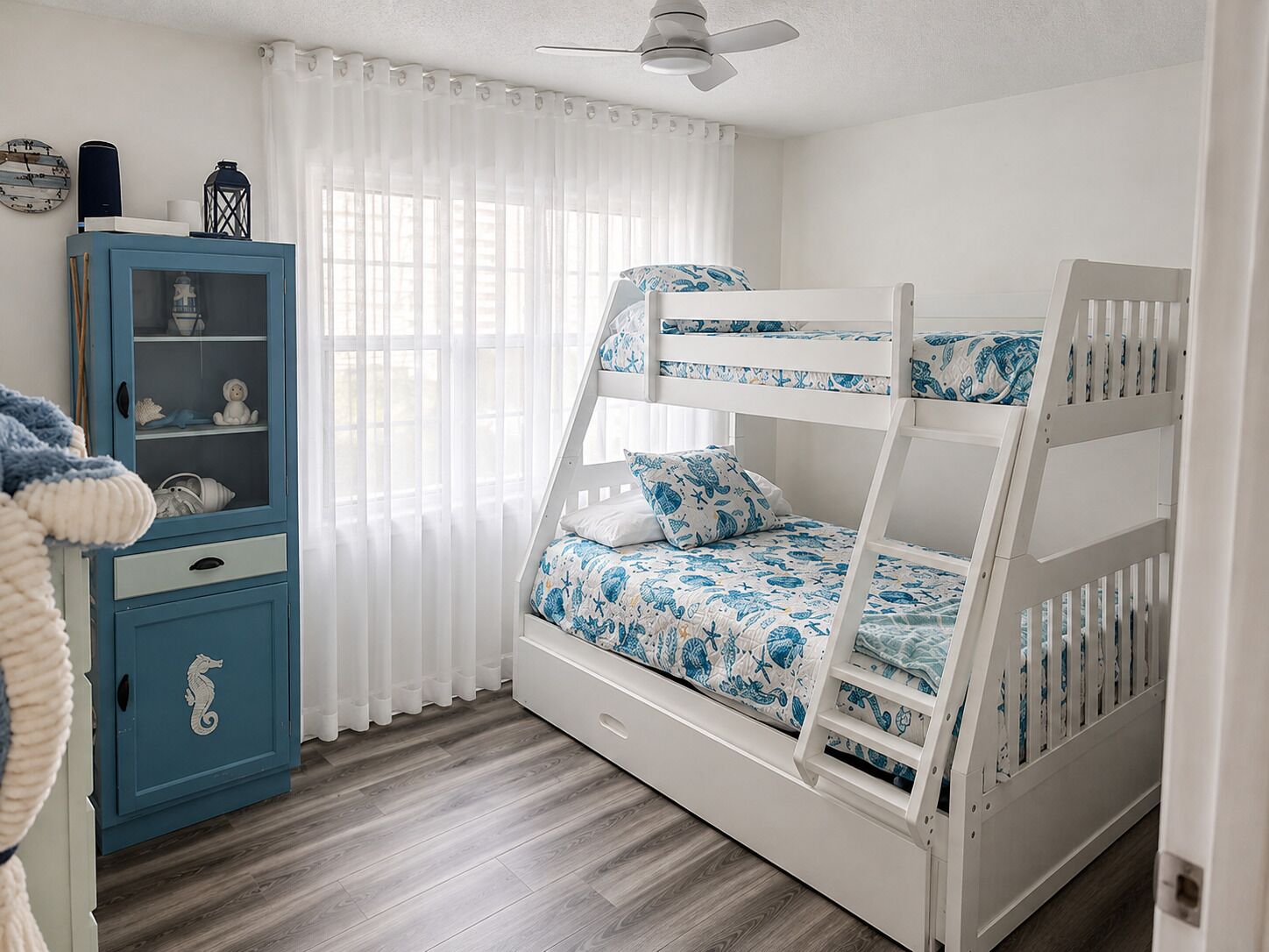 A bunk bed with a white frame and blue bedding, a blue cabinet with glass doors, and a white ceiling fan.