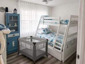 A child's bunk bed with a blue and white nautical theme, a blue cabinet with decorative items, and a grey playpen in a room with wooden flooring and white walls.