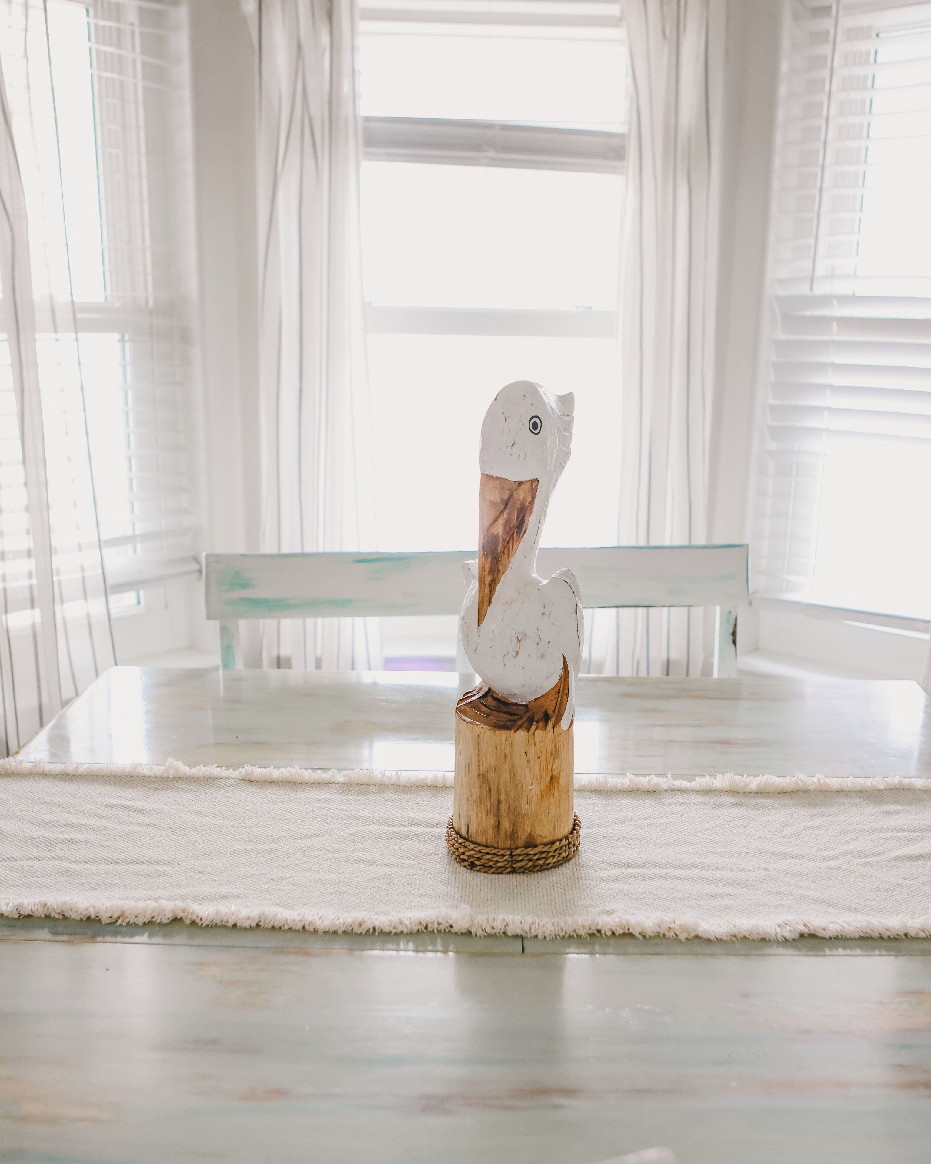 A white bird sculpture on a wooden base in a bright room with a glass table and white curtains.