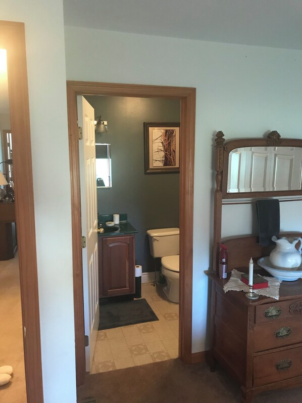 A narrow hallway leading to a bathroom with a toilet, sink, and framed artwork. A wooden dresser with drawers and a pitcher is visible in the foreground.