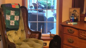 A room with a green upholstered chair, a wooden dresser, and a window with a view of outdoor furniture and greenery.