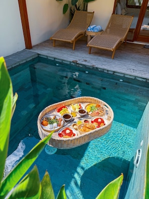 A poolside area with a wicker table holding a variety of food items, two wicker lounge chairs, and a potted plant.