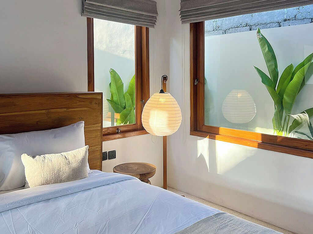 Ein Schlafzimmer mit einem Holz-Kopfteil, einem Bett mit weißer Bettwäsche, einem Nachttisch mit Lampe und großen Fenstern mit Milchglas und Grünpflanzen.
