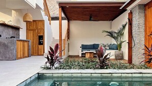 Een modern buitenzwembad met een houten pergola, een zithoek met een bank en salontafel, en een keuken met houten kastjes.