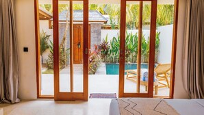 Une chambre dotée d’une porte-fenêtre coulissante donnant sur une zone avec piscine, d’un lit recouvert d’un couvre-lit à motifs et d’une petite table avec une chaise.