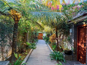 A wooden pathway leading to a building with a wooden door, surrounded by lush greenery and tropical plants.