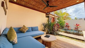 Een overdekt terras met een plafondventilator, een bank met kussens, een ronde tafel en uitzicht op een aangelegde tuin.
