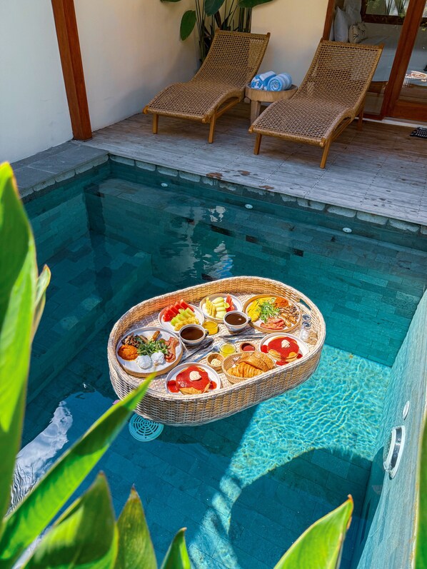 A poolside area with a wicker table holding a variety of food items, two wicker lounge chairs, and a potted plant.