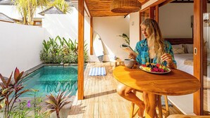 A person dining at a wooden table with a plate of fruit, near a pool with plants and a surfboard. - Muzeum Villas: 1 Bedroom, 1 Bathroom, Wi-Fi, Kitchen, Private Pool (Bali)