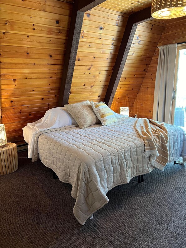 Une pièce chaleureuse, avec des murs en bois, comprenant un lit, une petite table d’appoint et une fenêtre agrémentée de rideaux.