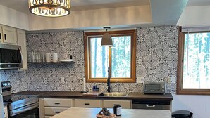 A modern kitchen with a central island, two wicker chairs, a chandelier, and patterned wallpaper. - New! Quiet cabin in the pines at Custer State Park (Custer)