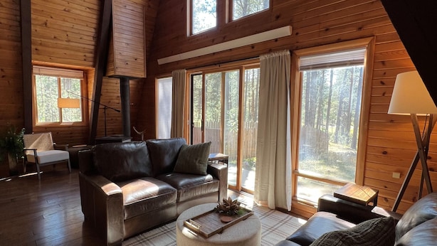 A spacious living room with a high ceiling and wooden walls, featuring a sofa, a coffee table, and large windows offering a view of the outdoors. - New! Quiet cabin in the pines at Custer State Park (Custer)