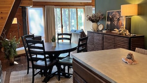 A cozy kitchen with a wooden table, chairs, and a large island. There is a window with curtains, a chandelier, and a clock on the wall. - New! Quiet cabin in the pines at Custer State Park (Custer)