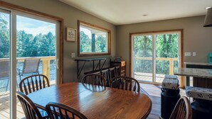 Comedor con mesa y sillas de madera, ventanas amplias con vista a árboles y una zona de cocina con mostrador y pileta.