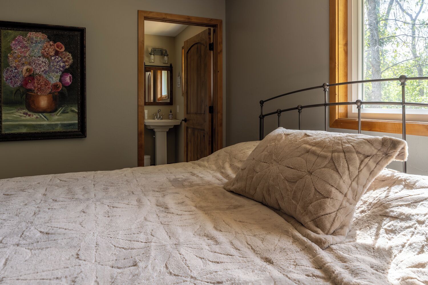 Un dormitorio con cama, ventana, un cuadro y un baño visible a través de una puerta abierta.