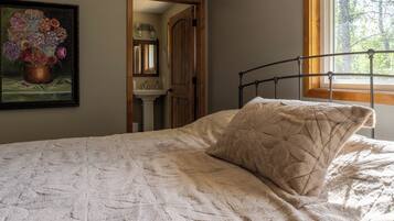 Un dormitorio con cama, ventana, un cuadro y un baño visible a través de una puerta abierta.