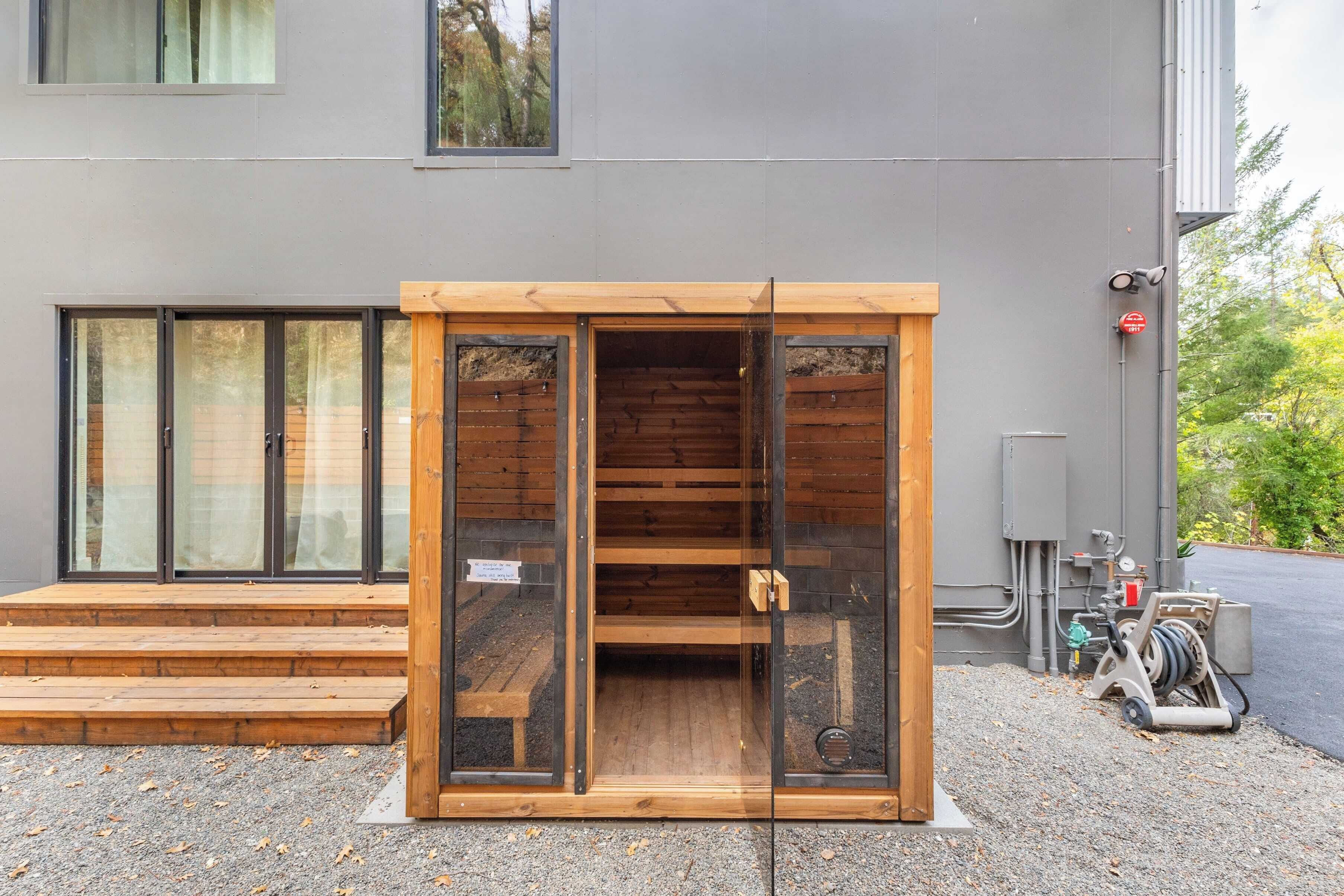 Ein modernes Gebäude mit Holzkonstruktion im Vordergrund, möglicherweise eine Sauna oder ein Lagerraum, und eine graue Außenwand mit Fenstern sowie einem Hydranten.